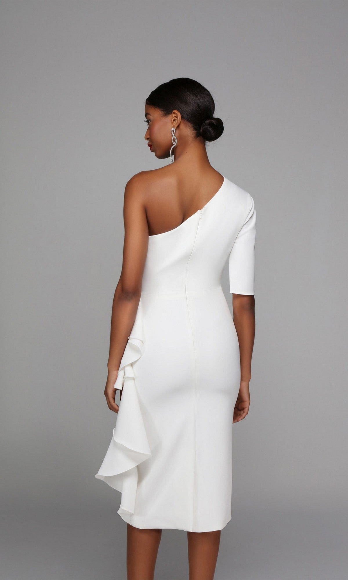 A woman with a low bun stands facing away, wearing the Alyce 70006 knee length semi-formal one-shoulder dress in white with a ruffle detail, set against a plain grey background.